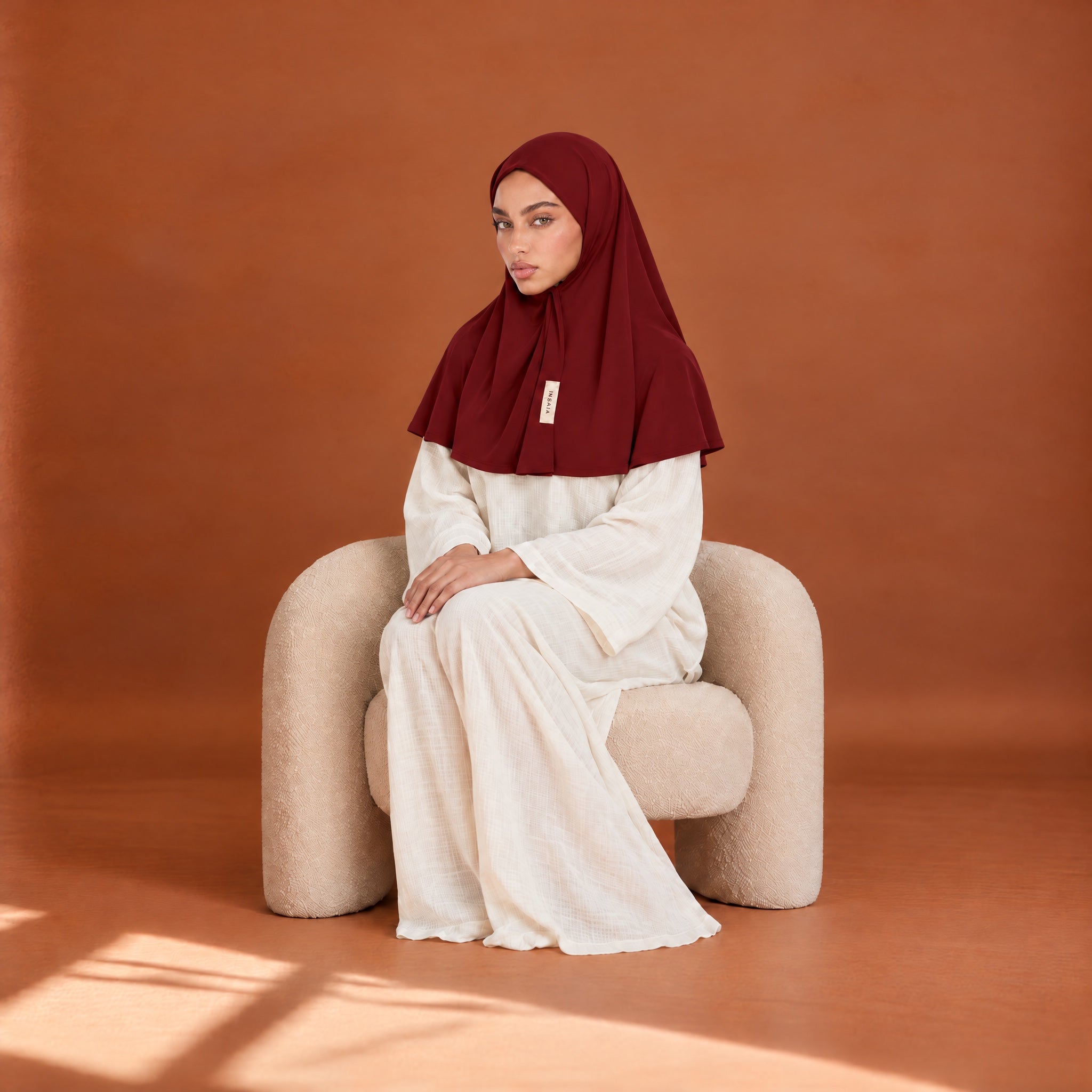Model wearing the INSAÏA Ruby jersey scarf, seated in a cream armchair against a terracotta wall