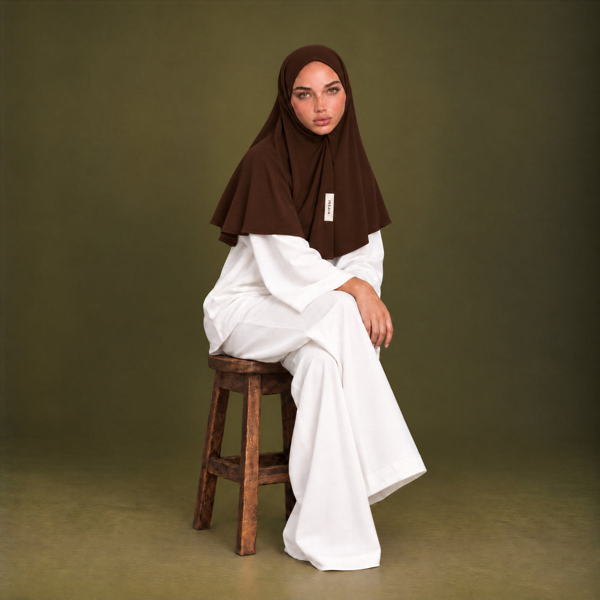 Model in the INSAÏA Dark Brown jersey scarf and a white two-piece set, seated on a wooden stool against an olive backdrop