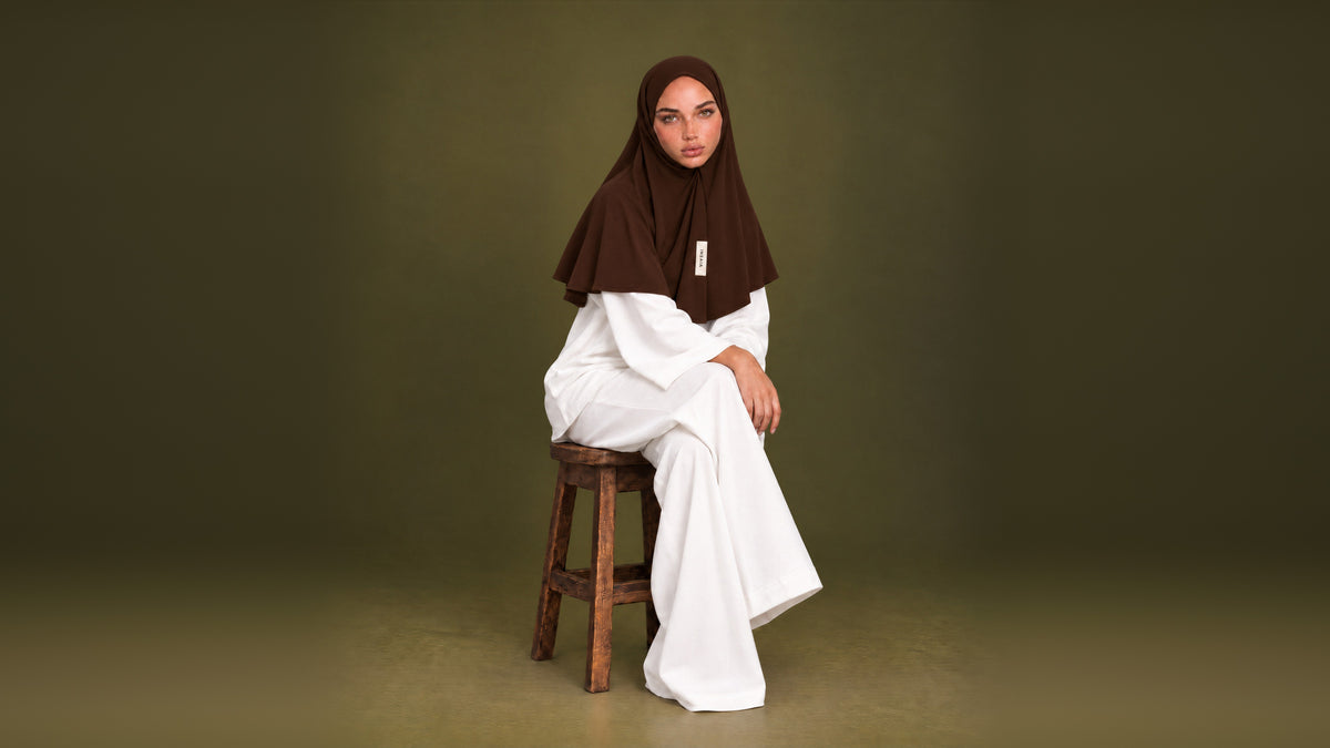 INSAÏA model in a dark brown jersey hijab seated on a wooden stool