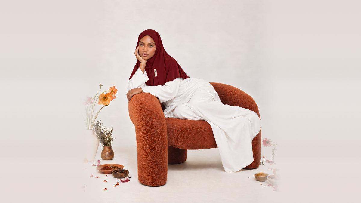 Model in the INSAÏA Ruby jersey scarf and white outfit, reclining in a terracotta armchair with flowers and spices